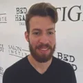 A bearded man with short, styled hair stands in front of a wall with Bed Head and Salon Institute logos.