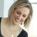 Smiling woman with short blonde hair wearing a black top and gold necklace, looking downward.