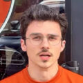 A young man with short brown hair, glasses, and a mustache stands in front of a glass door, wearing an orange shirt.
