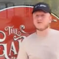 A person in a light shirt and black cap stands in front of a red sign that reads