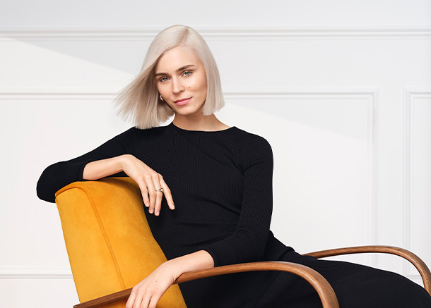 Woman with straight platinum blonde bob, wearing a black dress, sits on a yellow chair against a white paneled wall.