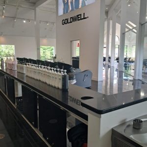 Modern hair salon interior with a row of hair product bottles on a counter and a large Goldwell advertisement on the wall.