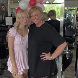 Two women standing and smiling in a salon, with pink and white balloons in the background.