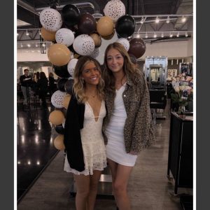 Two young women pose in front of a black, white, and gold balloon display inside a modern salon.
