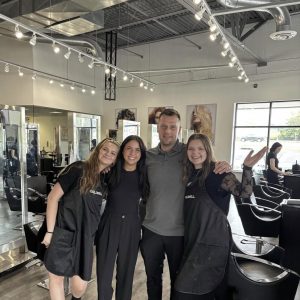 Four people pose together and smile inside a modern hair salon with black chairs and bright overhead lighting.