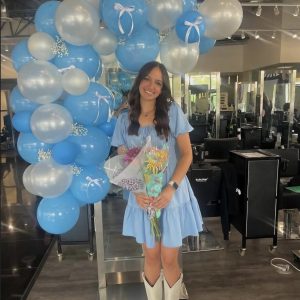 Woman in a light blue dress and white boots holding flowers, standing by blue and white balloons in a salon.