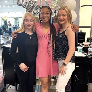 Three women pose together indoors in front of a celebratory balloon display with "congrats" in silver letters.