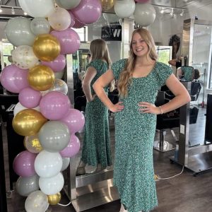 A woman in a green floral dress stands by a balloon arch, smiling, with her reflection visible in a mirror behind her.