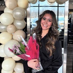 A young woman in a black outfit holds a bouquet of flowers, standing in front of white balloons and smiling at the camera.