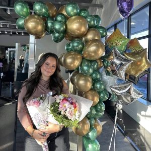 A woman holding a bouquet of flowers stands in front of green and gold balloons and "Congrats" balloons.