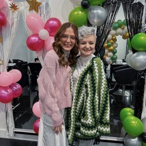 Two people smiling and posing together at a party with balloon decorations and tinsel curtains in the background.
