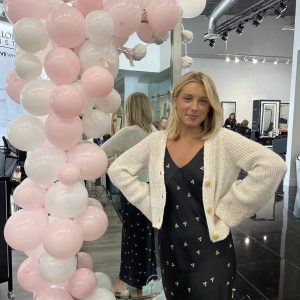 A woman in a black dress and white cardigan stands beside a pink and white balloon display in a modern salon.