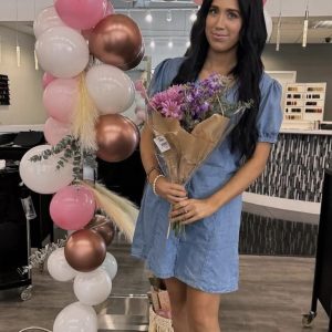Woman in a denim dress holding a bouquet of flowers stands in front of a pink and white balloon arch indoors.
