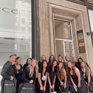 A group of people in black outfits stand outside a building entrance, holding branded tote bags and smiling at the camera.