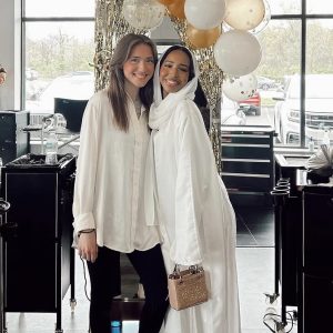 Two women standing and smiling indoors, with balloons and gold streamers in the background.