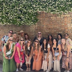 A group of people pose outdoors in front of a brick wall with blooming white flowers overhead on a sunny day.
