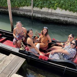 Five women sit and smile in a gondola on a canal, with greenery and a stone wall in the background.