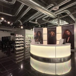 A modern salon reception desk with illuminated front, large posters, and product shelves in a sleek, industrial space.