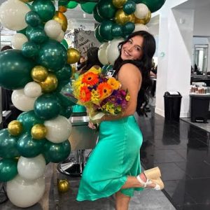 Woman in green dress poses with a bouquet of flowers in front of a green, white, and gold balloon arch indoors.