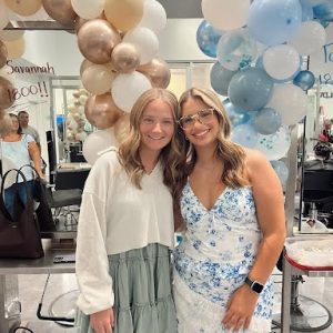 Two women smiling and posing inside a decorated room with blue and gold balloon arches in the background.