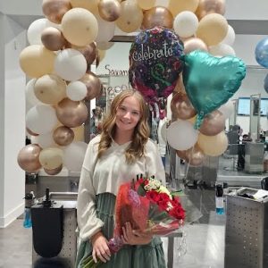 Young woman holding flowers and balloons stands in front of a balloon arch at an indoor celebration event.