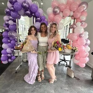 Three women stand smiling between purple and pink balloon arches, holding flowers inside a brightly lit room.