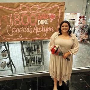 A woman in a white dress holds flowers, standing by a sign reading "1800 and done, Congrats Aizlynn" in a store.