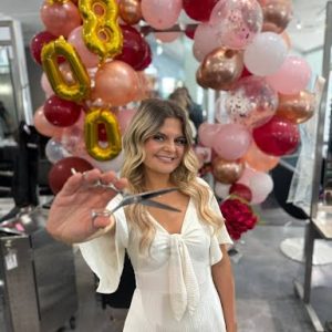 Woman in white dress holding scissors, standing in front of pink and gold balloon arch with "18" balloons.
