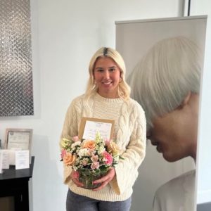 Woman holding a bouquet and certificate stands indoors in front of a poster with a close-up of a person’s hair.