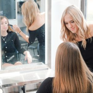 A hairstylist styles a client's hair in a salon, both facing a large mirror with bright natural light.