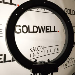 A ring light stands in front of a backdrop featuring the Goldwell and Salon Institute logos.