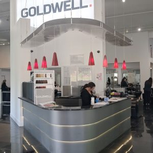 Reception desk at a modern salon with a Goldwell sign, red pendant lights, and a staff member working behind the counter.