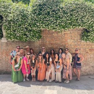 A group of people pose in front of a brick wall with green foliage and white flowers on a sunny day.