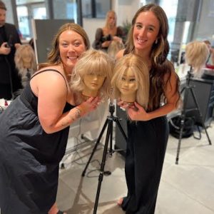 Two women in a salon smile and pose holding mannequin heads with styled blonde wigs on tripods.