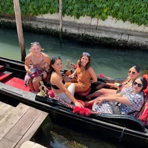 Five women sit in a gondola on a canal, with greenery on the wall in the background and sunlight overhead.