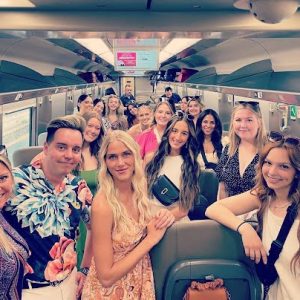 A group of people pose and smile for a photo inside a train carriage, with seats and windows visible.