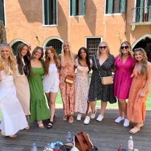 Nine women in colorful dresses stand side by side on a wooden dock by a canal with buildings in the background.