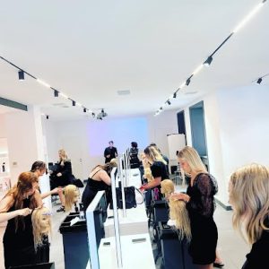 A group of people practice hairstyling on mannequin heads at workstations in a brightly lit salon classroom.