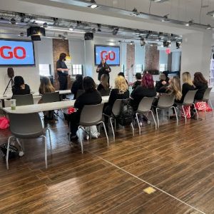 People seated at tables listen to two presenters standing in front of screens displaying the word "GO" in a modern room.