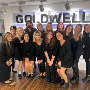 A group of thirteen people in black outfits pose and smile in front of a Goldwell sign indoors.