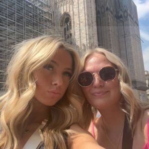 Two women pose for a selfie in front of the Milan Cathedral, with crowds and scaffolding visible in the background.